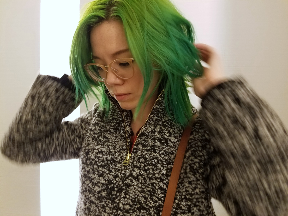 a photo of Alex King, in a black and white speckled coat, looking slightly downward and fluffing her green hair